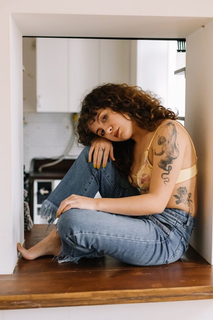 Curly-haired woman with tattoos sits in a cozy indoor space wearing denim.